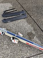 Full view of one ski with Tyrolia bindings attached and two black Yakima ski rack parts on pavement.