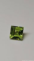 Close-up photo of a bright apple green peridot gemstone, showing the square step cut and reflective polished surfaces.