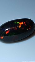 Close-up photo of the black Ethiopian opal cabochon showing bright multi-color flashes in red, green, and orange hues.