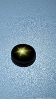 Oval cabochon black star sapphire showing full 6-ray star flash under lighting, smooth polished surface.
