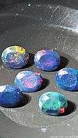Close-up photo of six oval faceted black opal gemstones showing vibrant multicolor flashes with a dark background.