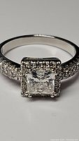 Close-up front view of ring displaying square princess cut center moissanite and accent stones on band.