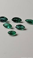 Six marquis cut emerald gemstones arranged on white surface, showing deep green color and natural inclusions.