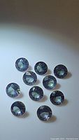 Clear image showing eleven round mystic topaz gemstones with diamond cut facets and iridescent color play on a plain background.