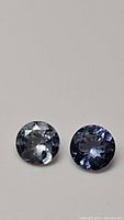 Two round polished natural tanzanite gemstones placed side by side, showing differences in shade from light blue-gray to deeper purplish-blue.