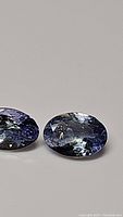 Close-up photo of two oval cut tanzanite stones with visible internal reflections and violet-blue color, lying side by side on a flat surface.
