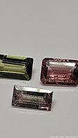 Three emerald cut tourmaline gemstones in olive green, pink, and light pinkish-purple hues shown on white background with visible facets.