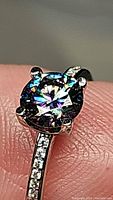 Close-up of the ring showing the round brilliant cut rainbow colored Moissanite stone with green, blue, and purple flashes under light.