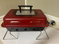 Red portable rectangular metal camping BBQ with black folding legs and a black handle on top. Vent holes visible on sides, and a control knob on the right side for gas regulation.