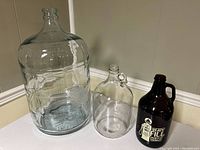 Three glass containers on a corner surface: large clear glass carboy, medium clear glass carboy with label, and brown glass growler with handle and printed label.