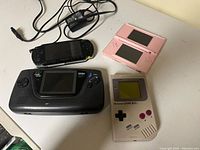 Four handheld consoles arranged on a white table: Nintendo GameBoy original, Sega Game Gear, Sony PSP, and pink Nintendo DS Lite. PSP power adapter also visible.