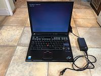 Open IBM ThinkPad T40 laptop showing screen and power adapter connected, placed on floor tiles.