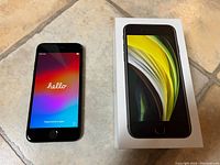 Front view of iPhone SE powered on displaying 'Hello' screen next to its retail box