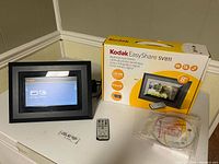 Kodak EasyShare SV811 digital picture frame powered on showing touchscreen interface, placed near remote control, disc and box on table.