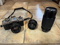 Minolta XG-M camera body with attached 50mm f/1.7 lens, plus two additional lenses (28mm and 80-200mm) laid on tile floor