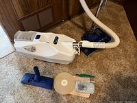 Full view of Miracle Mate Platinum vacuum cleaner main unit with attached hose, Sebo blue power head, blue vacuum attachment tool, and beige vacuum bags along with small extras.