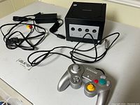 Nintendo GameCube console with controller, AC adapter, and AV cables laid out on table, cables visible and controller connected.