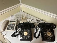 Two black Northern Telecom rotary dial phones and one white Radio Shack phone with large push buttons on table, cords and handset showing.