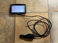 TomTom XL GPS showing language selection on its touchscreen with car charger connected, on tiled floor.