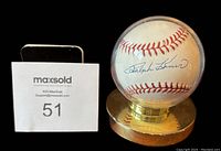 Ralph Kiner signed baseball displayed in a custom gold-tone stand with clear acrylic cover. Signature visible on the white leather baseball under protective case.