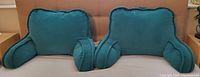 Pair of two teal backrest bed pillows with arms and pockets, set on a bed.