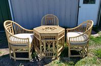 Full set of 3 rattan chairs with cushions arranged around octagonal rattan table outdoors on grass with some gravel.