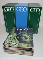 Three Geo magazine holders (two green, one blue) containing multiple issues of Geo magazines with one Geo magazine on top showing the Statue of Liberty on cover.