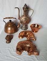 Full view of copper teapot, ewer, bowl, wall mount and four copper molds arranged on white background