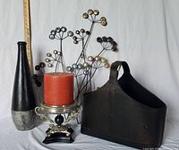 Front view of the decor lot showing the 3-wick candle on a decorative holder, black and silver vase, dark brown magazine holder, and metal wall decor in the background.