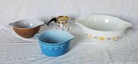 Four items arranged in a row on white cloth: three Pyrex bowls (white large with floral pattern, medium brown, small blue with pattern) and one glass carafe with metal lid and plastic handle.