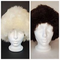 Pair of two Cossack hats on white mannequin heads. The white dyed Tuscan lamb fur hat is fluffy and medium size. The brown dyed lamb fur hat has ear flap-like points and a slightly smaller size.