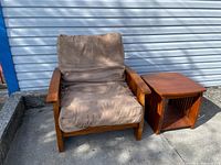 Armchair with brown cushions and wooden frame, next to matching wooden side table on concrete outside a building with white siding.
