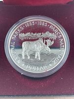 Close-up of the commemorative silver dollar coin showing the moose in water with forest and mountains, and inscriptions commemorating the National Parks Centennial.