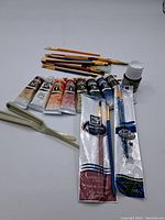 Lot arranged with oil paint tubes, various paintbrushes, and a small bottle on white background.