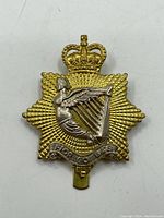 Front view of WWII Irish Regiment of Canada cap badge showing gold-tone starburst base, silver-tone winged figure on harp, crown detail, and regiment banner.