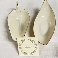 Two leaf shaped small porcelain dishes in ivory or cream with gold trim; one with handle and embossed leaf veins; Lenox brand card placed between them for scale and identification.