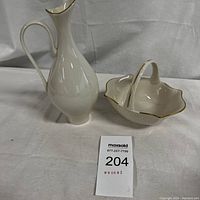 Two Lenox porcelain pieces on white fabric: a vase with a handle and a gold rim, and a handled decorative dish with a scalloped edge and gold trim.