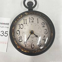 Front angle of the decorative retro clock showing the dial, hands, and wear on the black plastic frame with gold decorative elements.