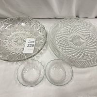Four clear pressed glass salad bowls displayed on a white cloth background. Two larger bowls have distinct pressed glass patterns, two smaller matching bowls with scalloped edges.