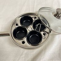 Stainless steel egg poacher pan with four black nonstick cups, pan side visible, cups fit into recessed sections, lid beside pan