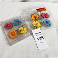 Two sets of four colorful hibiscus flower table weights in plastic packaging on white background.