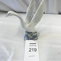 Front view of the white porcelain swan figurine with wings spread, positioned on a blue wave-like base with a MaxSold lot tag in front.