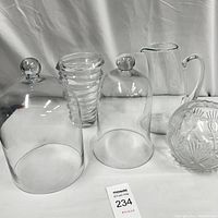 Five clear glass kitchen decor pieces including two tall bell jars with lids, a pitcher, textured vase, and cut glass bowl