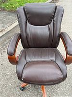 Front view of a brown leather Lazboy office chair showing padded armrests with wooden accents, seat cushion with wear including creases and a small damaged area on the front left.