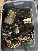 Top view of storage bin with wireless keyboard, Belkin case, USB charger, wireless mouse, and various tangled cables.