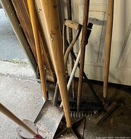 Collection of garden and construction tools arranged leaning against a wall in a garage. Tools include shovels, rakes, and brooms with wooden handles showing signs of wear and rust on metal parts.
