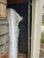 Photo of a large black metal highway sign frame stored upright next to a plastic-covered item and a wooden barrel. Shows the side of the sign with rails for letter attachment.