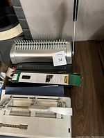 Metal comb binding machine with black handle, white hole punch, blue binder, and packaging box on wooden table.