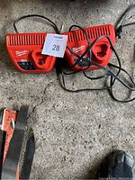 Photo showing two red Milwaukee M12 18V battery chargers placed on concrete floor with cables intertwined.