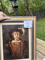 Full view of painting and frame, showing a young woman in historical attire and a large hat with feather, outdoors on wooden surface.